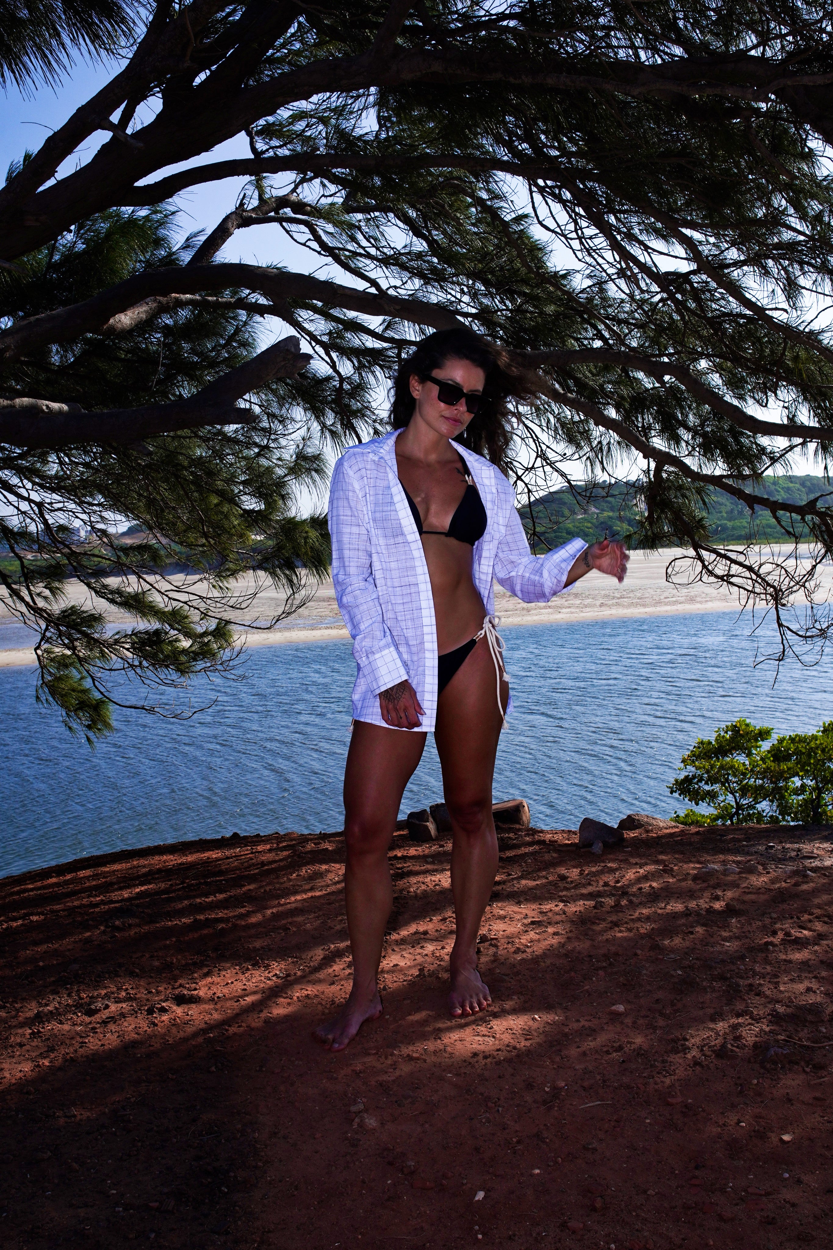 Woman in a white cardigan and black bikini standing by a body of water with trees in the background