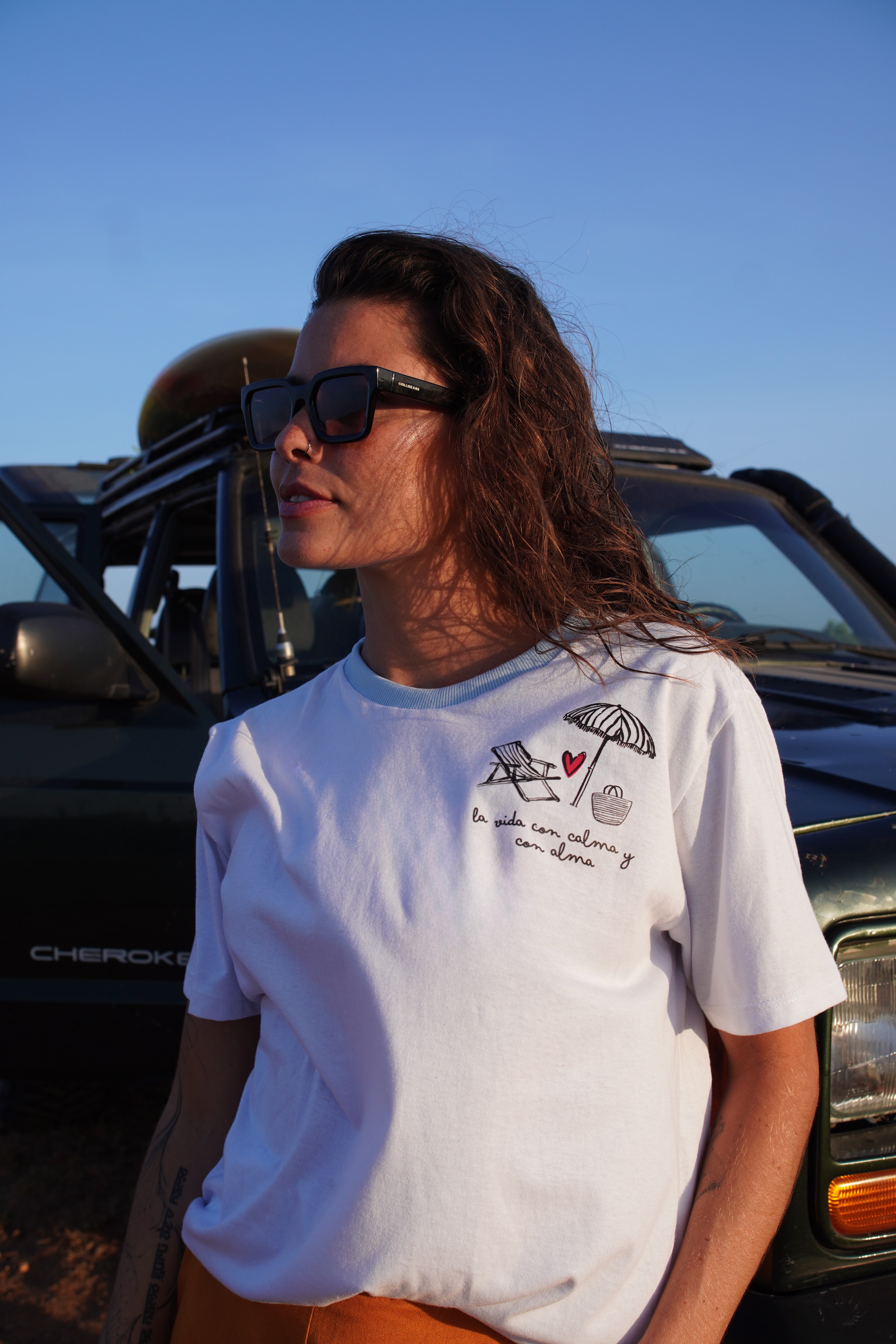 Woman wearing a white t-shirt with graphics and text, standing in front of a green vehicle.
White graphic T-shirt with minimalist embroidered details, styled for a relaxed summer look — blending casual sophistication with timeless resort lifestyle.