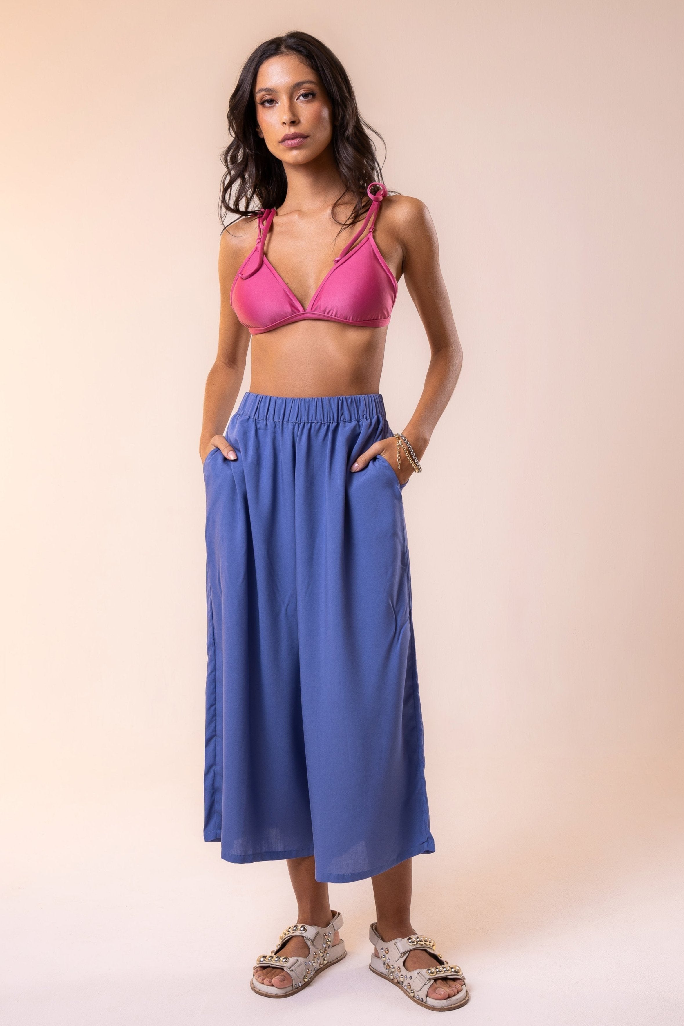A woman standing, wearing blue pants with a wide, elastic waistband, and two side pockets. She is also wearing a pink swimsuit top.
Woman wearing a pink bikini top and high-waisted blue flowy pants from Hoy Swim, showcasing a minimalist resort look that blends tropical elegance with effortless comfort.