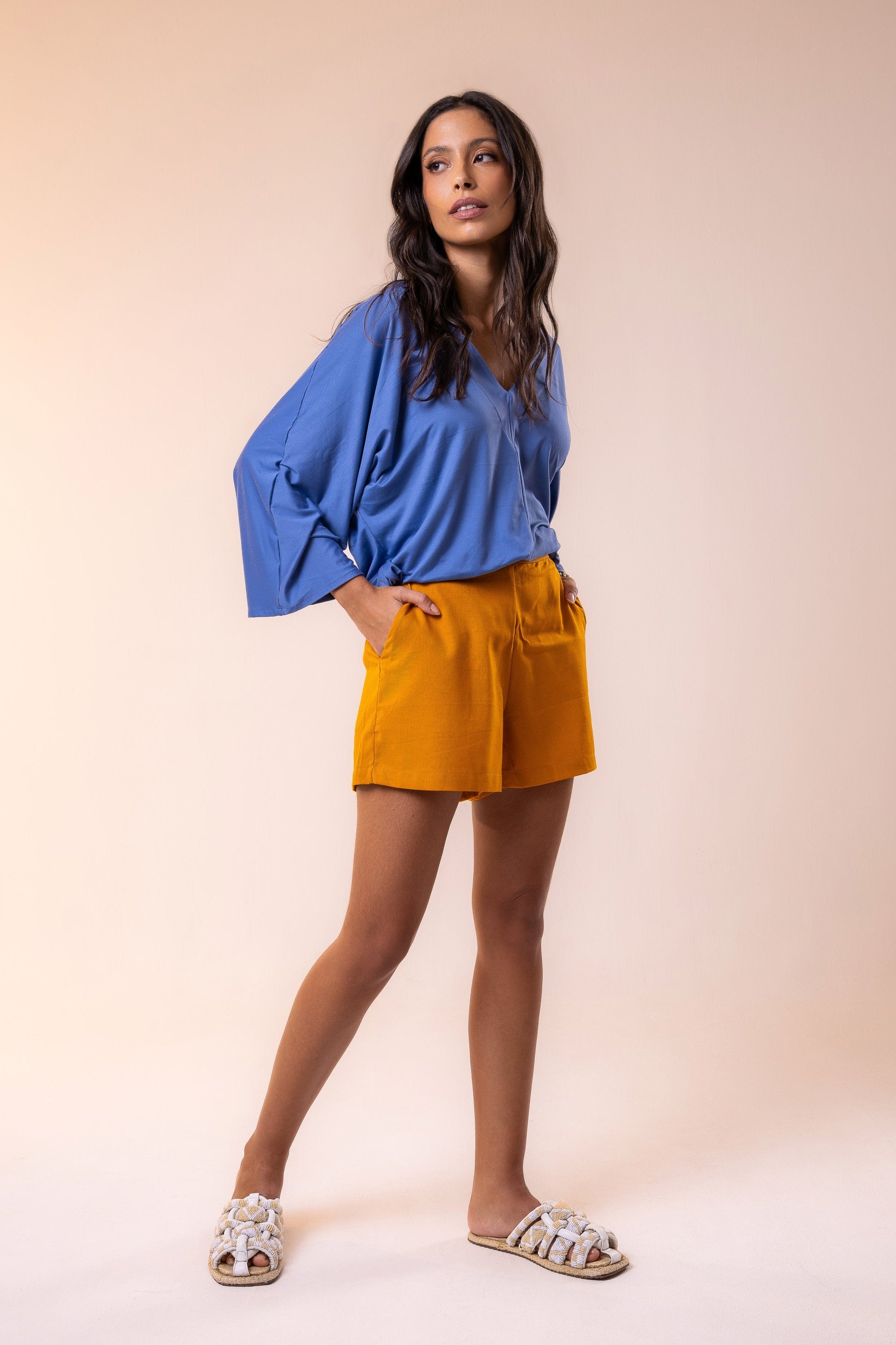A female model wearing a blue blouse with a loose fit and a pair of orange shorts, standing against a beige background.
Chic resort look featuring a relaxed viscose blouse in ocean blue paired with tailored ochre shorts — a modern combination of effortless style and tropical elegance.