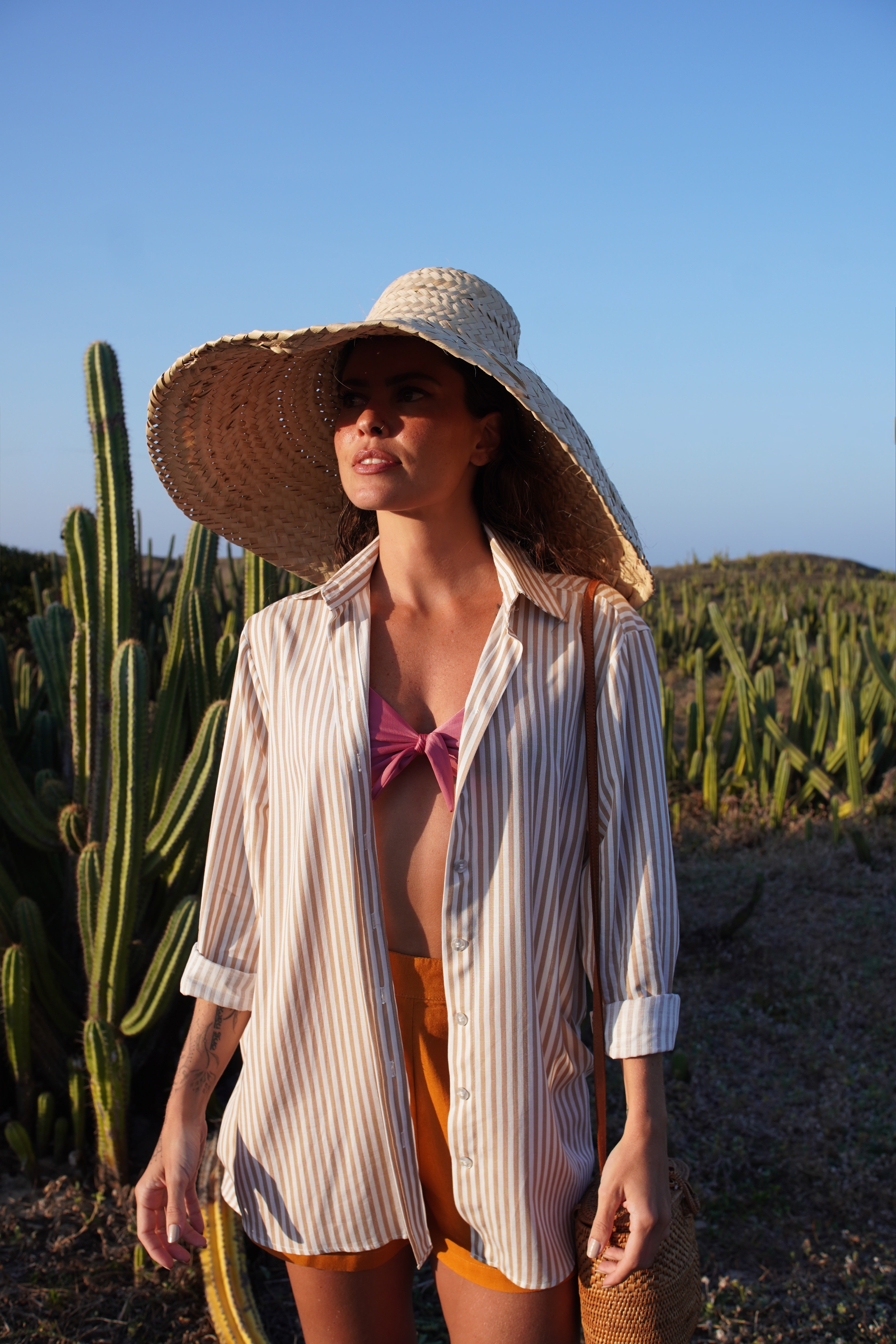 Striped lightweight resort shirt styled over a pink bikini top with tailored shorts and a wide sun hat — an elevated beachside look blending relaxed elegance and tropical sophistication