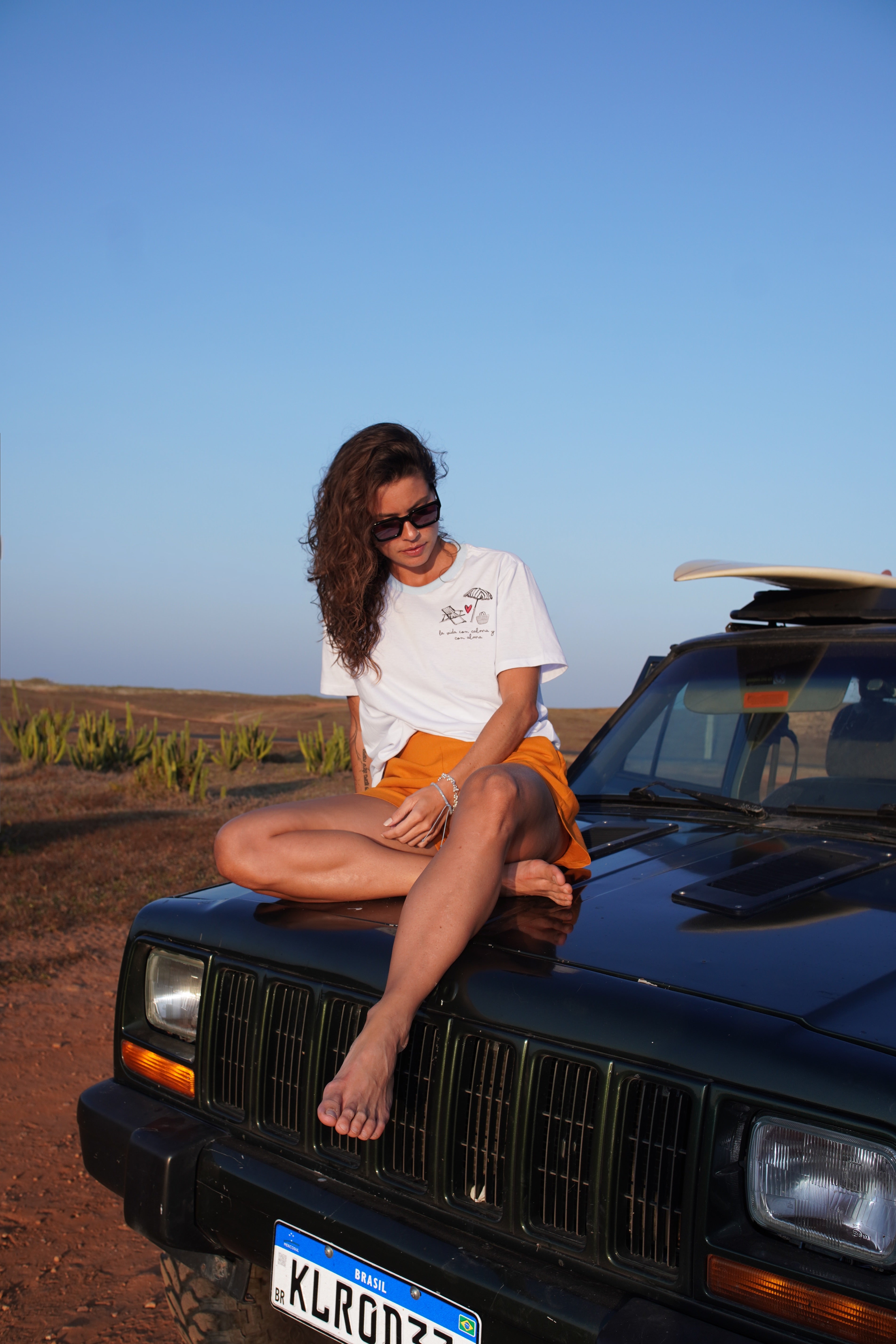 Woman sitting on a green vehicle with a clear blue sky in the background. Casual comfort meets timeless coastal style.