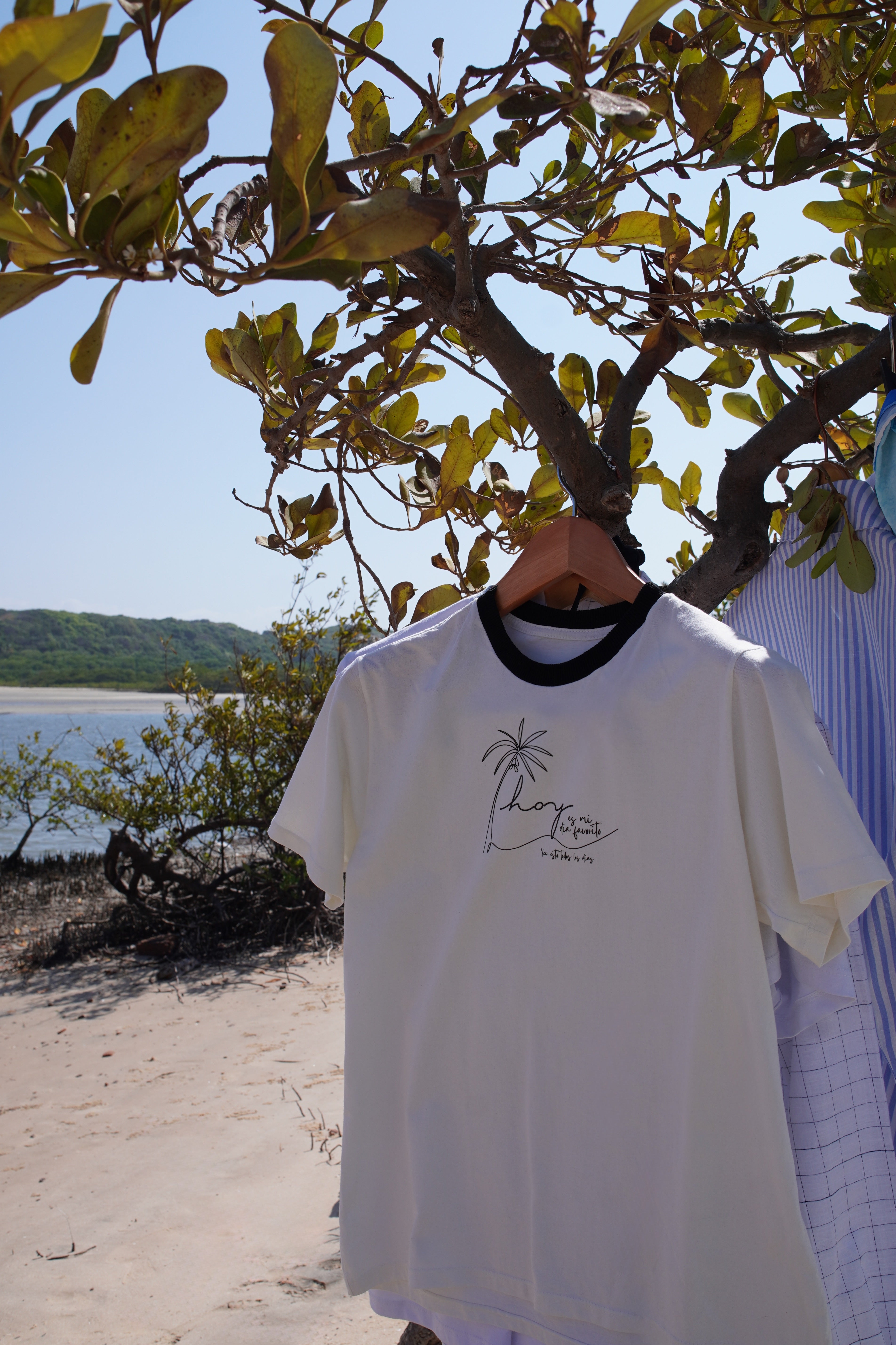 White Hoy Swim t-shirt with a palm tree print hanging on a tree branch by the beach, reflecting the brand’s natural, effortless, and tropical resort style from Brazil.