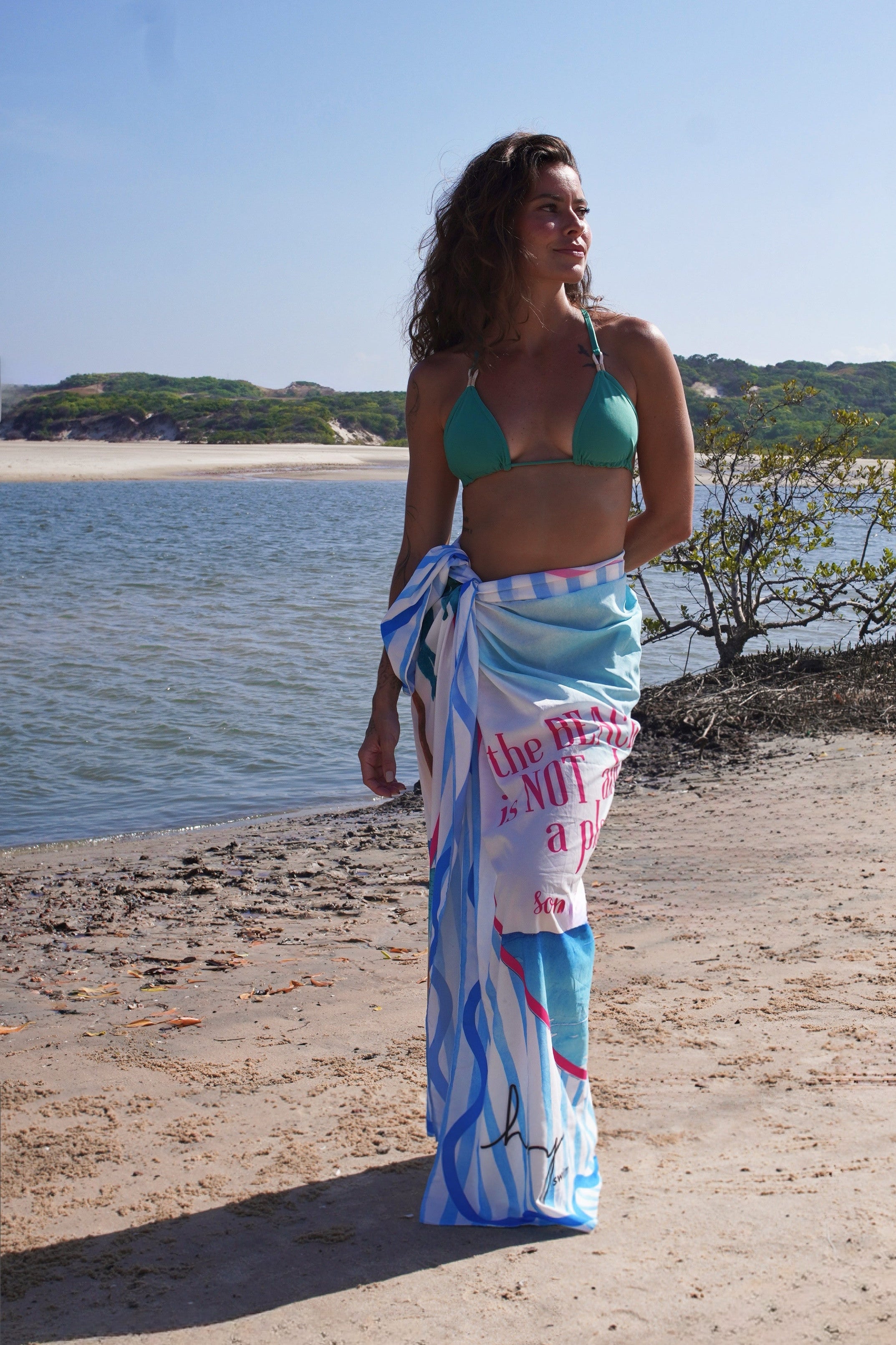 Woman in a green Hoy Swim bikini wears a blue and white printed pareo with pink details by the seaside, embodying the brand’s elegant, tropical, and effortless aesthetic.